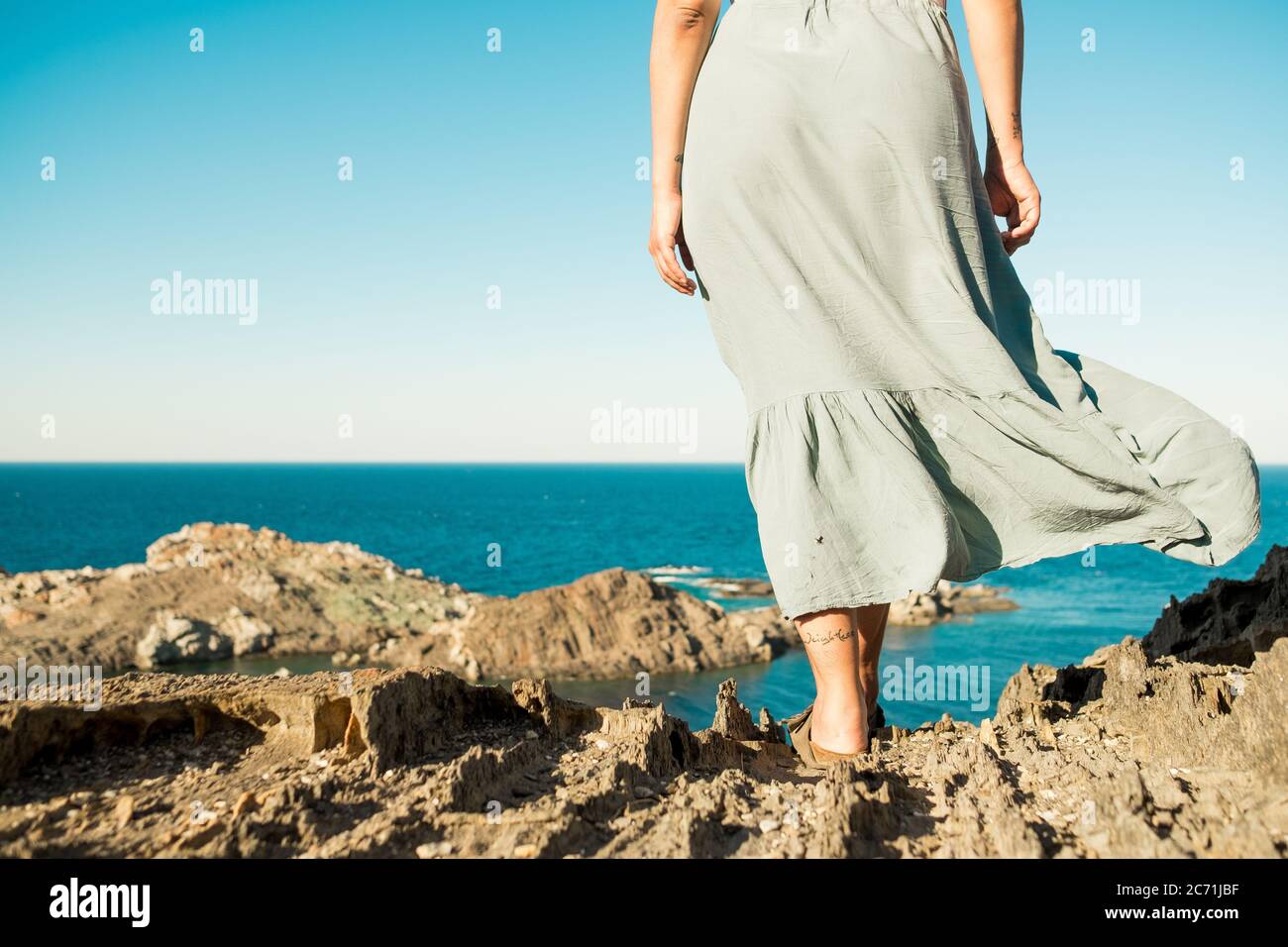 giovane e coraggiosa donna in piedi sulla cima della roccia in una giornata estiva guardando epico paesaggio marino con vestito in movimento nel vento, evocativo e stimolante vi schiena Foto Stock
