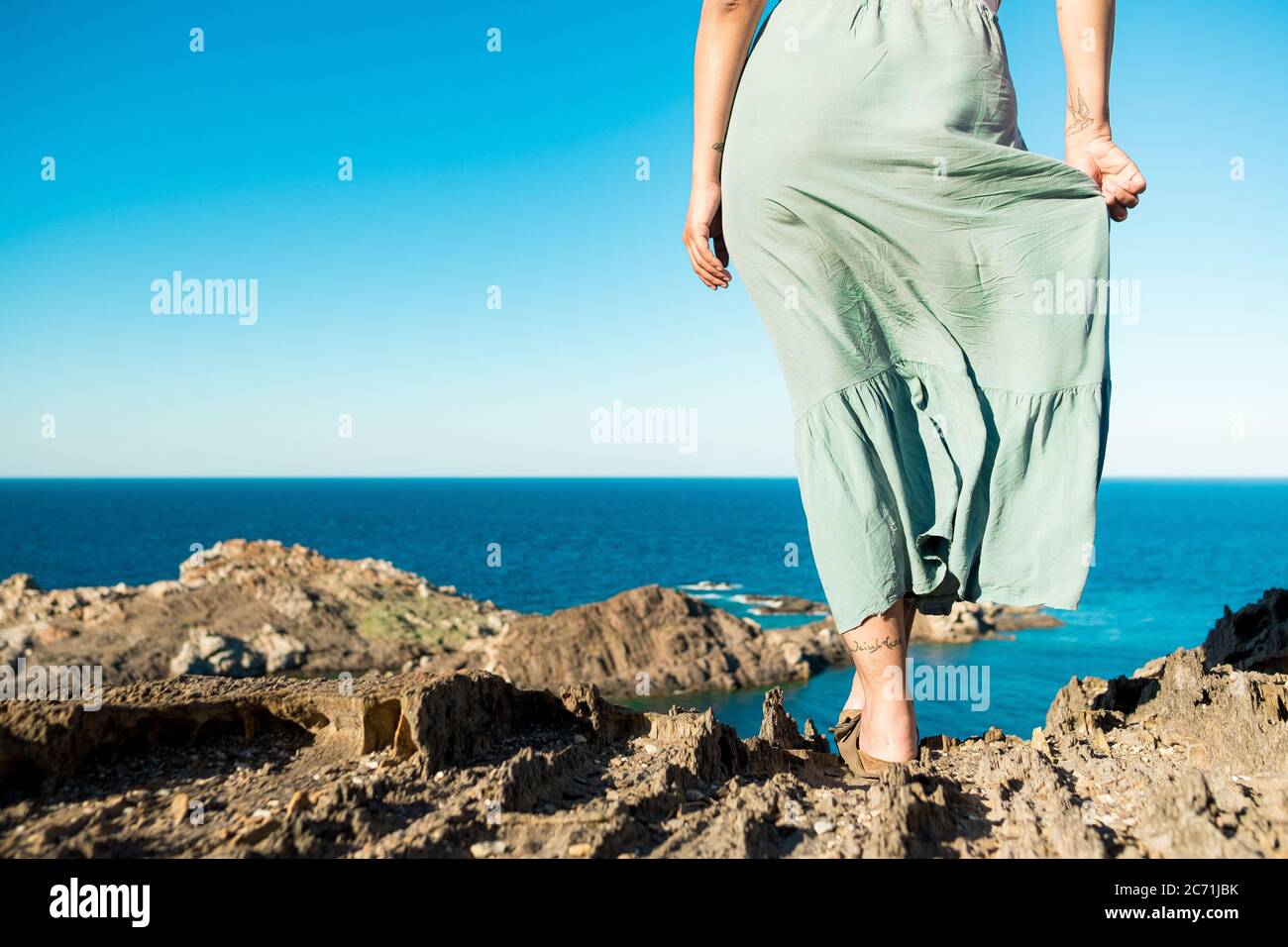 giovane e coraggiosa donna in piedi sulla cima della roccia in una giornata estiva guardando epico paesaggio marino con vestito in movimento nel vento, evocativo e stimolante vi schiena Foto Stock