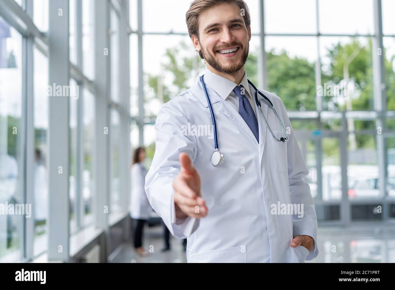 giovane medico che accoglie nel suo ufficio. medico uomo sorriso, tenere mano gesto di benvenuto con stetoscopio. Foto Stock