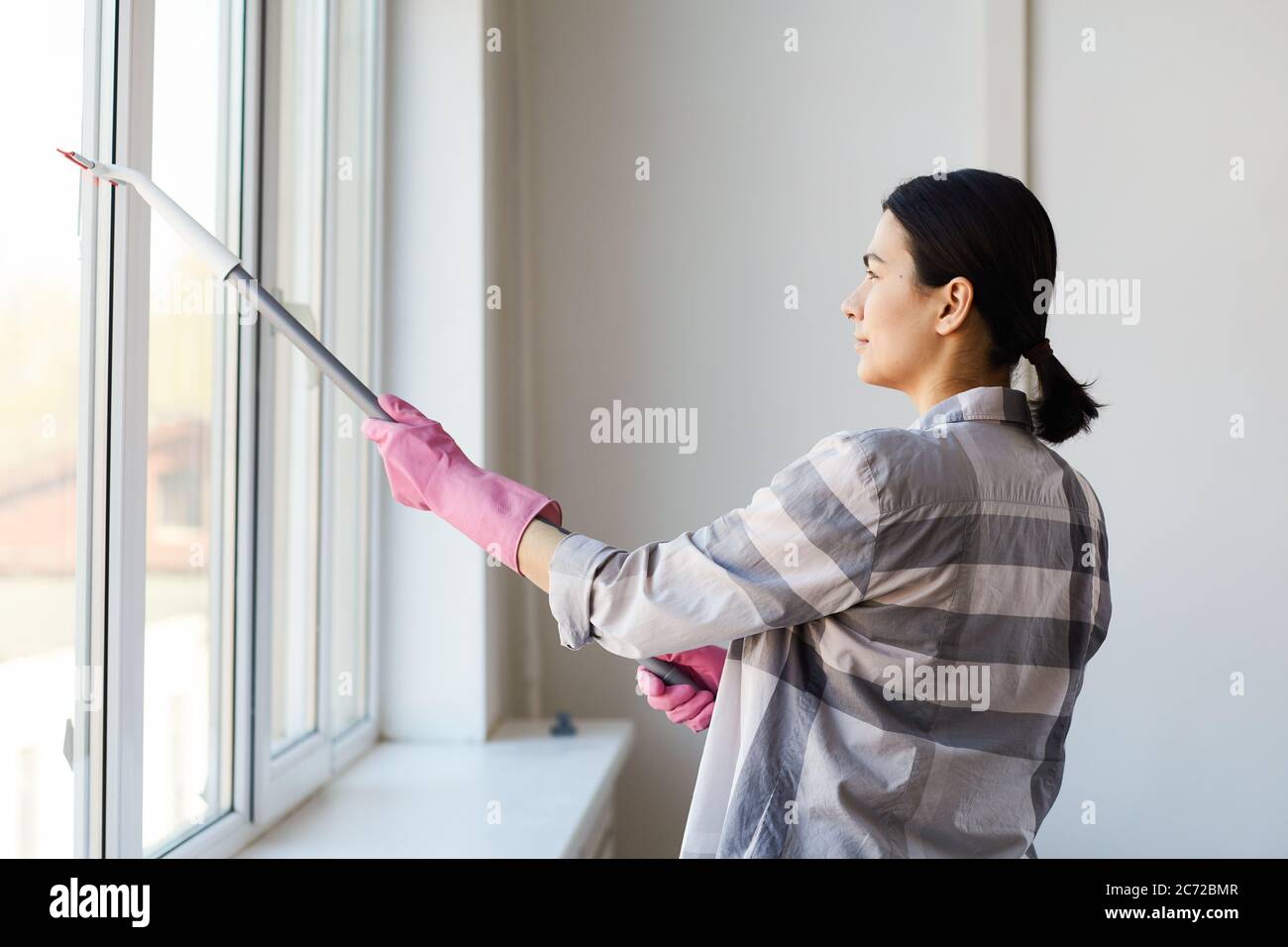 Giovane donna in guanti per pulire la finestra con la mop in ufficio Foto Stock