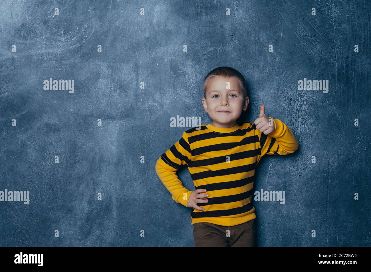 Ragazzo molto felice che fa ridere le dita. Il ragazzo si darà via in una giacca giallo-nero e pantaloni marroni che si pongono sullo sfondo della b Foto Stock