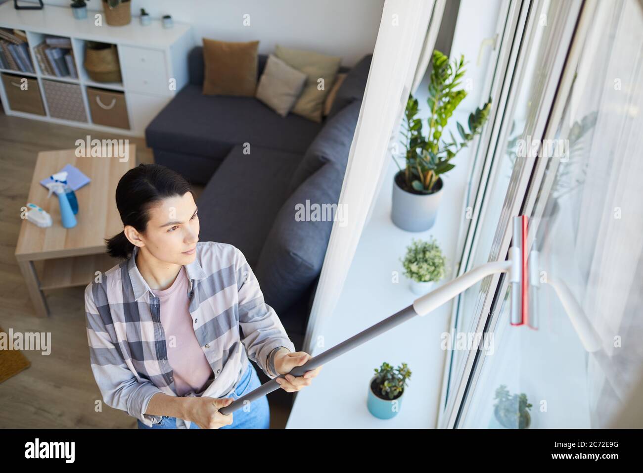 Giovane donna che usa la spugna e pulisce la finestra in camera a casa Foto Stock