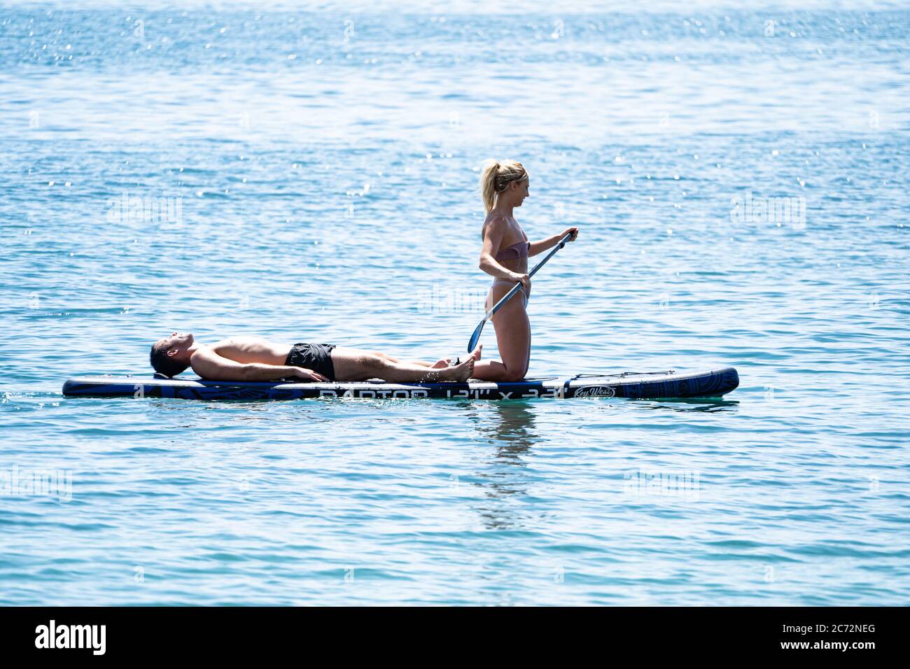 Una giovane coppia, uomo e donna, su un SUP (Stand up paddleboard), l'uomo sdraiato (addormentato?) E la donna si inginocchiò e si pagaiò dolcemente, Dorset, UK Foto Stock