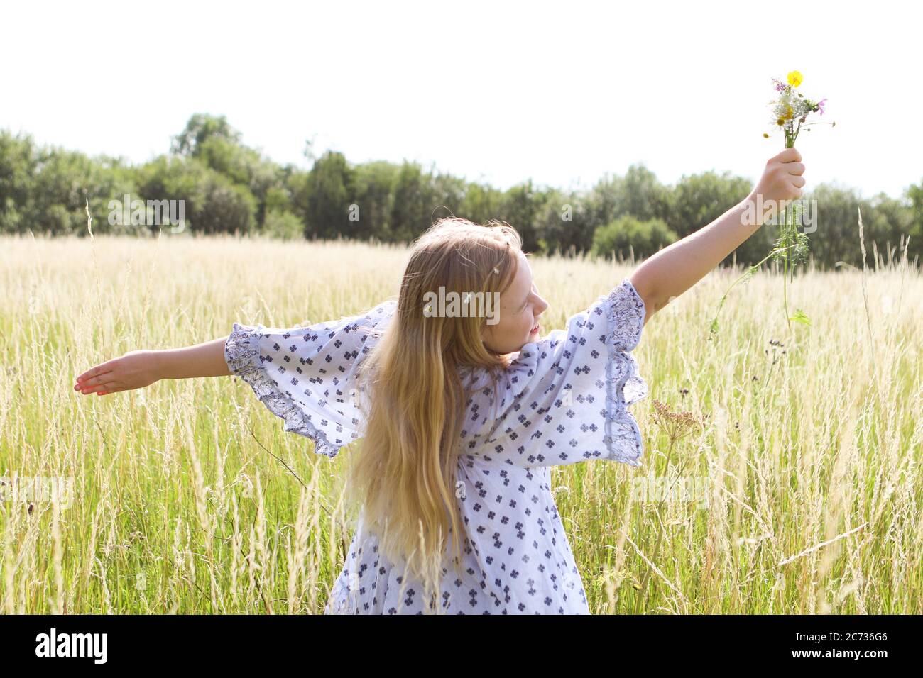 Una giovane ragazza hippy graziosa con catena daisy in capelli biondi tiene un poy di fiori selvatici con le braccia estese n un campo di pascolo in estate Foto Stock