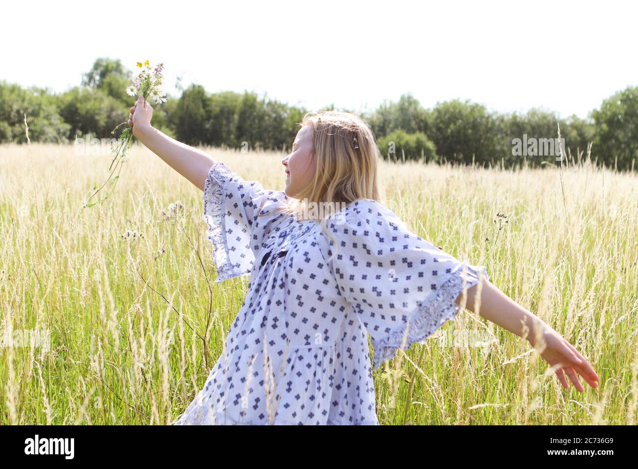 Una giovane ragazza hippy graziosa con catena daisy in capelli biondi tiene un poy di fiori selvatici con le braccia estese n un campo di pascolo in estate Foto Stock