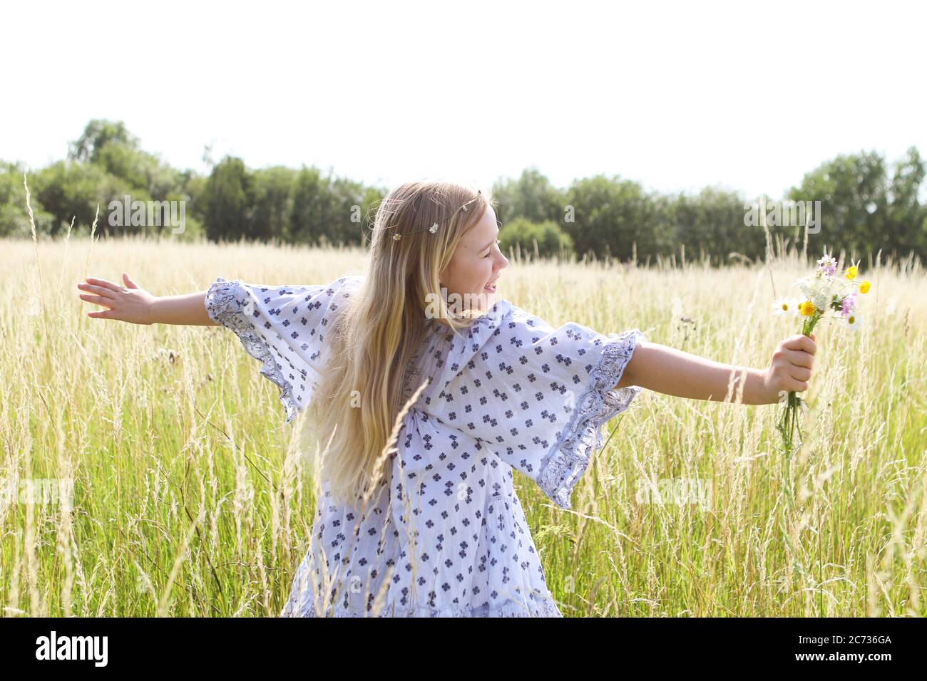 Una giovane ragazza hippy graziosa con catena daisy in capelli biondi tiene un poy di fiori selvatici con le braccia estese n un campo di pascolo in estate. Foto Stock