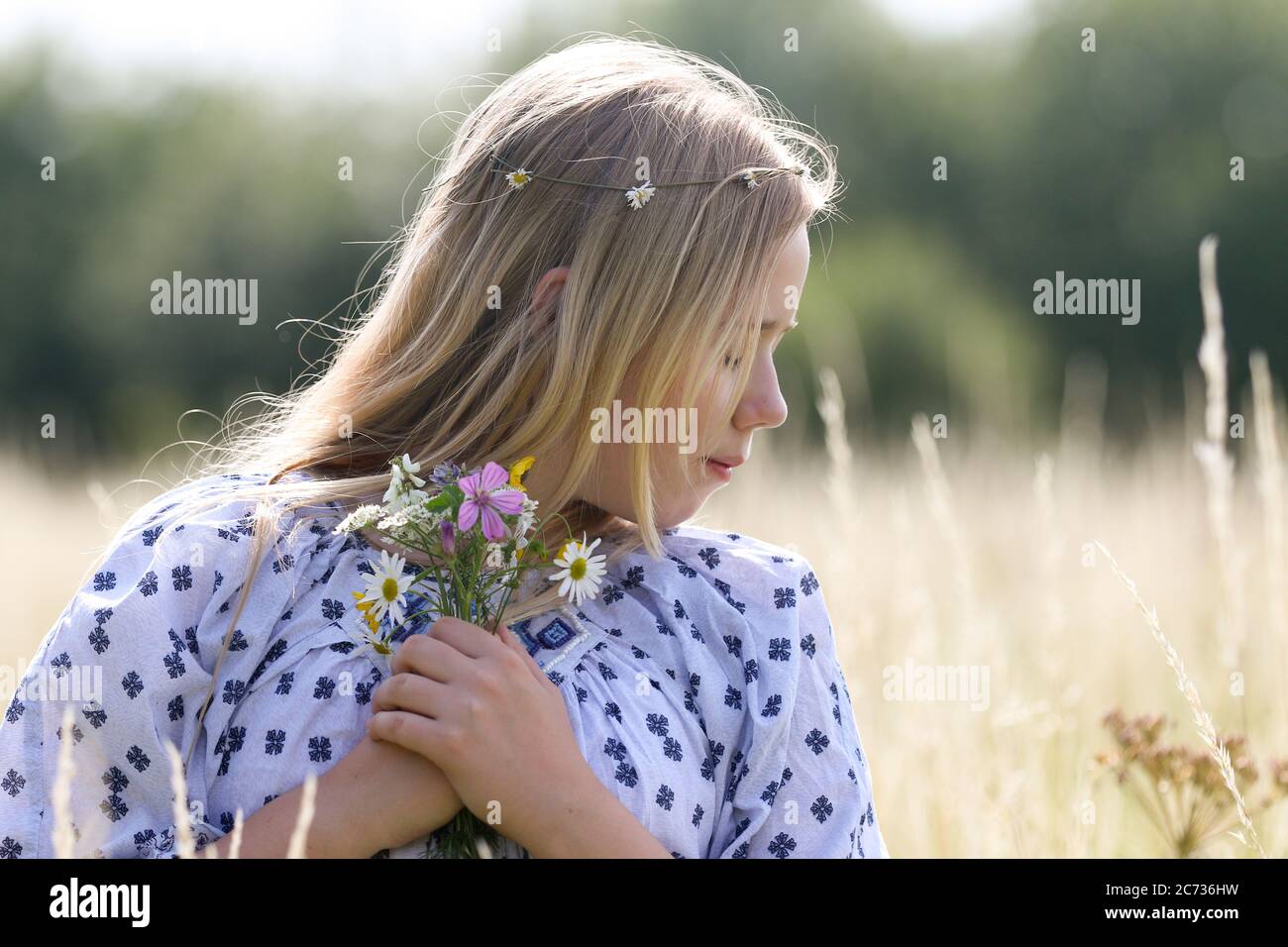 Una giovane ragazza hippy graziosa con una catena a margherita nei suoi capelli biondi tiene un poy dei fiori selvatici nel sole in un campo di pascolo in estate. Foto Stock