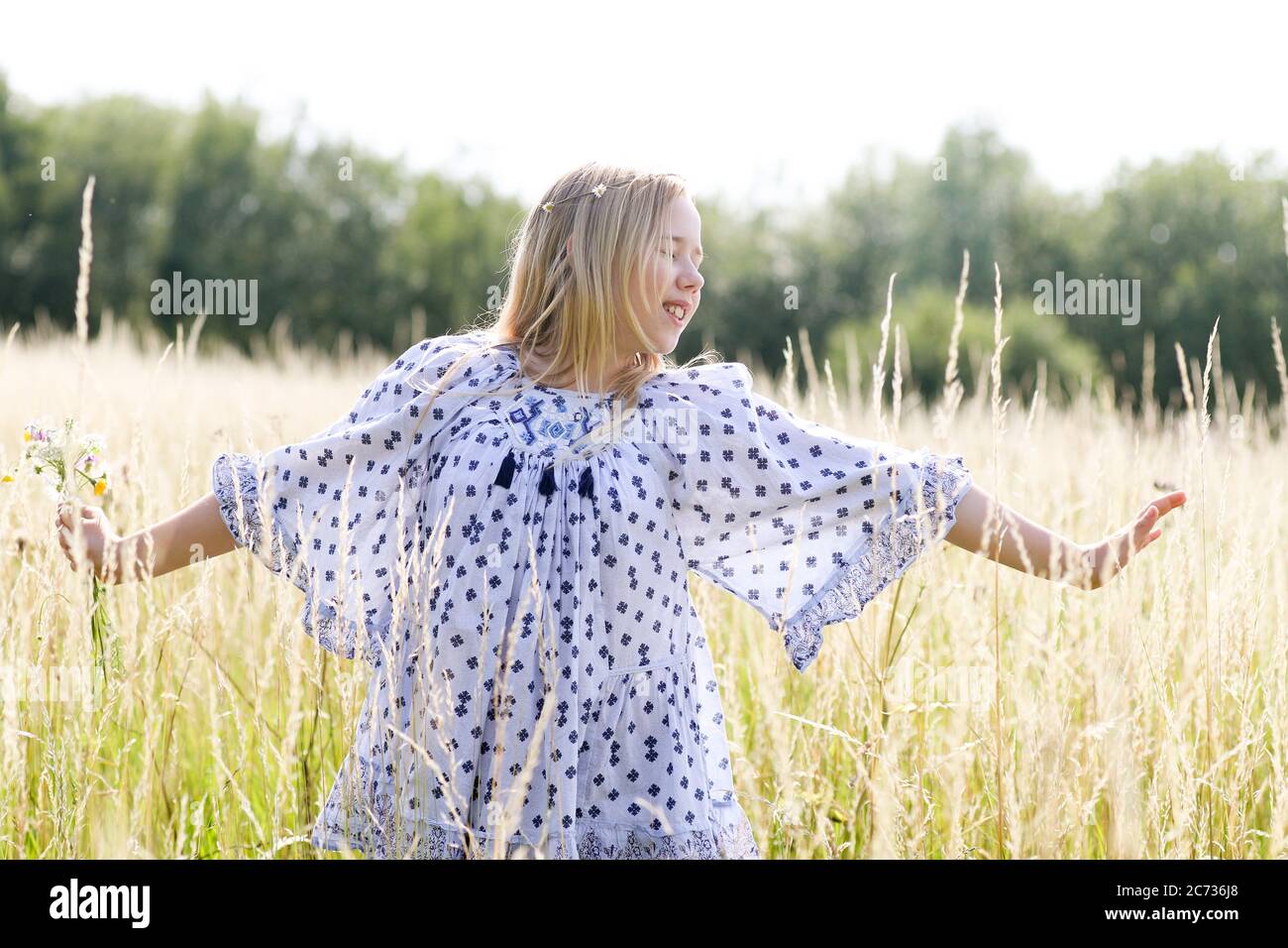 Una giovane ragazza hippy graziosa con catena daisy in capelli biondi tiene un poy di fiori selvatici con le braccia estese n un campo di pascolo in estate. Foto Stock