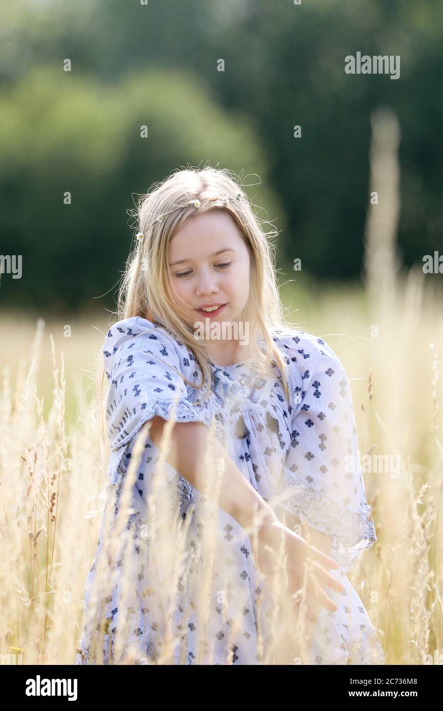 Una giovane ragazza hippy graziosa con una catena a margherita nei suoi capelli biondi cammina nell'erba lunga nel sole in un campo di pascolo in estate. Foto Stock