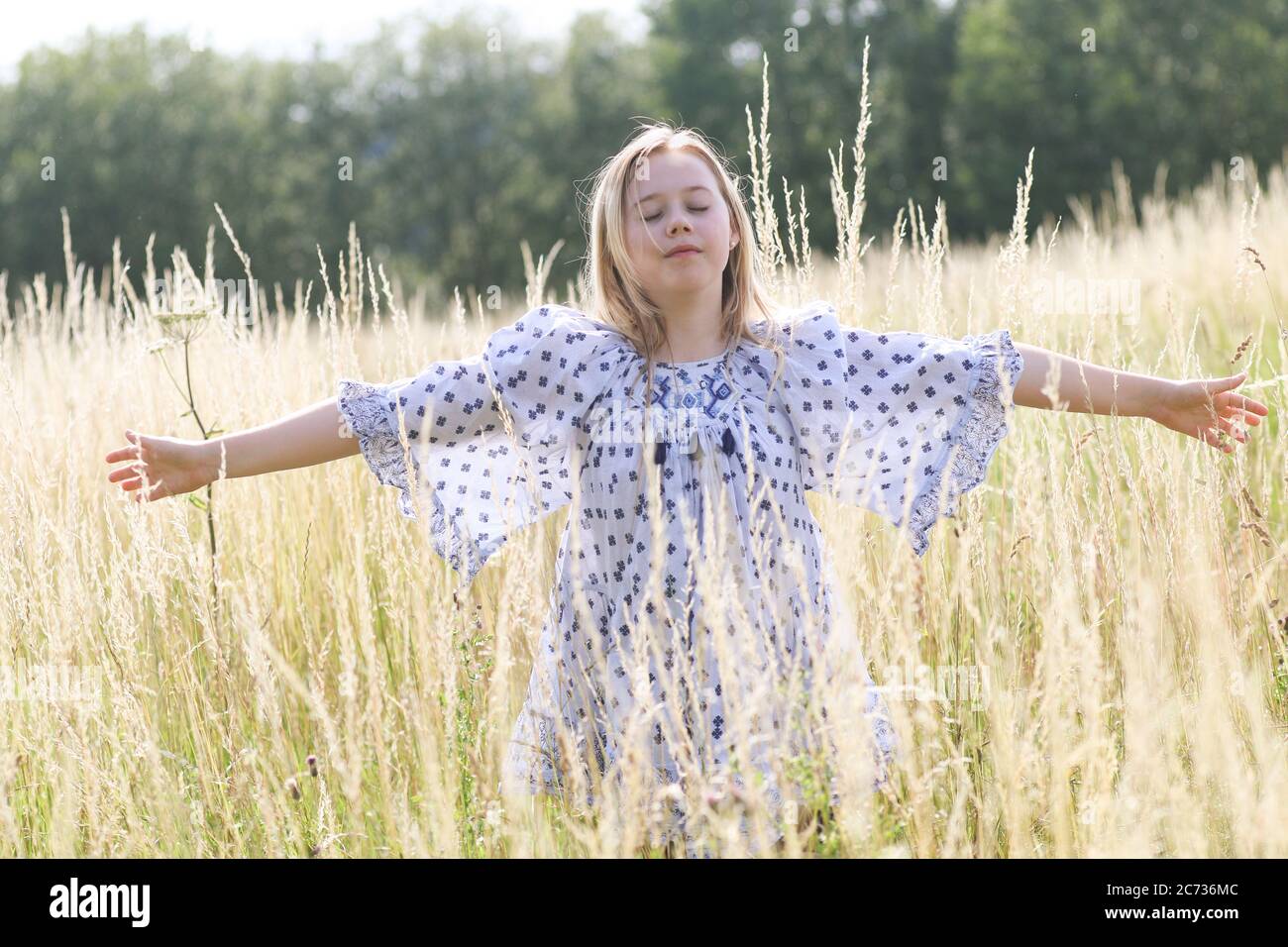 Una giovane ragazza hippy graziosa con i capelli biondi con le braccia estese nell'erba lunga di un campo di pascolo in estate. Foto Stock