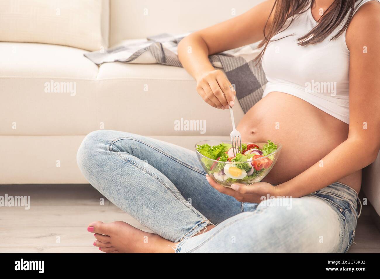 Donna che si aspetta un bambino mangia insalata da una ciotola di vetro seduta sul pavimento di un appartamento. Foto Stock