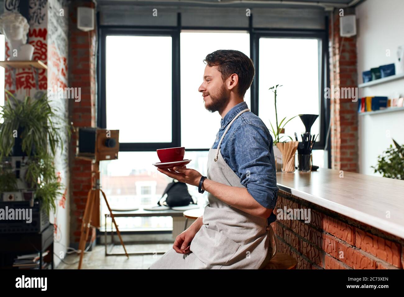 Ottimista barista maschile allegro, si siede vicino al bancone bar, tiene bella tazza con caldo gustoso americano, guarda da parte con viso positivo, sorridendo gentilmente, Foto Stock