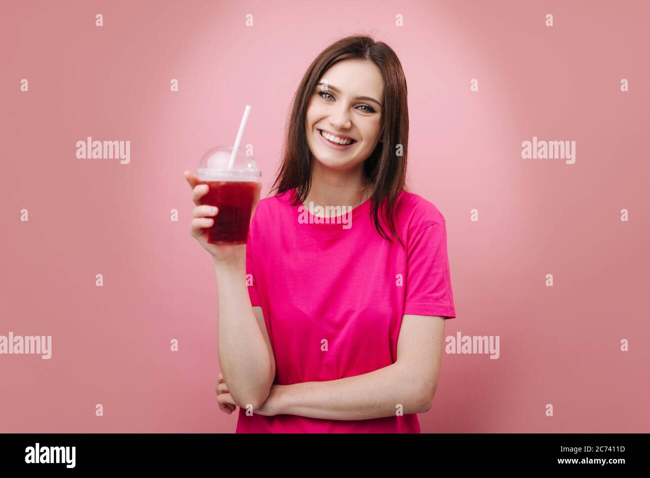 Ritratto di giovane ragazza allegra in t-shirt rosa con coppa di plastica con fresco cocktail freddo su sfondo rosa studio. Bella donna con h marrone Foto Stock