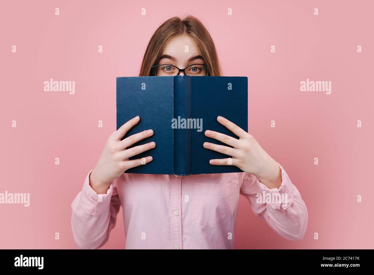 Donna bionda emozionale in occhiali nascondendo il suo volto dietro libro aperto su sfondo rosa. Bella ragazza in camicia casual che posa e guardando camer Foto Stock
