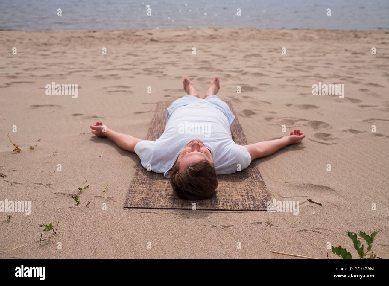 Giovane uomo che fa yoga su un lago in una giornata estiva, meditazione, posa relax, Shavasana o uomo morto asana Foto Stock