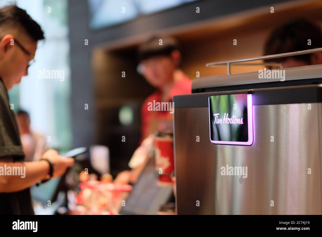 Il marchio canadese Tim Hortons apre il suo primo negozio in Cina. Primo piano sul logo lucido della macchina da caffè. Visione sfocata di impiegati e clienti Foto Stock