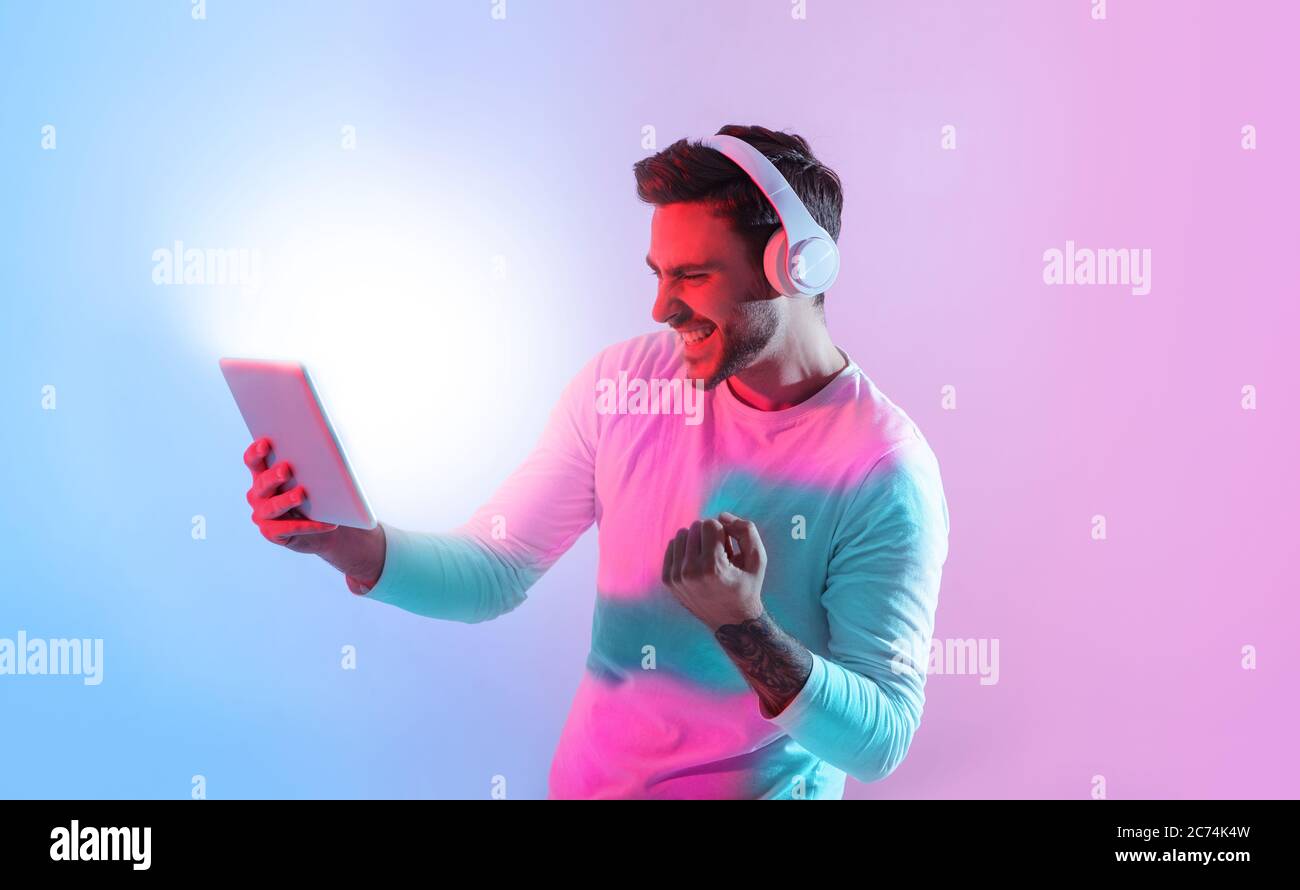 Vincere online. Il ragazzo millenario in cuffia celebra la vittoria, tenendo un tablet Foto Stock