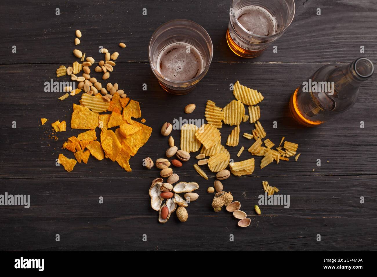 Su tavolo di legno scuro sono patatine, nachos, noci e pistacchi, nelle vicinanze sono due bicchieri di lager chiaro e bottiglia Foto Stock