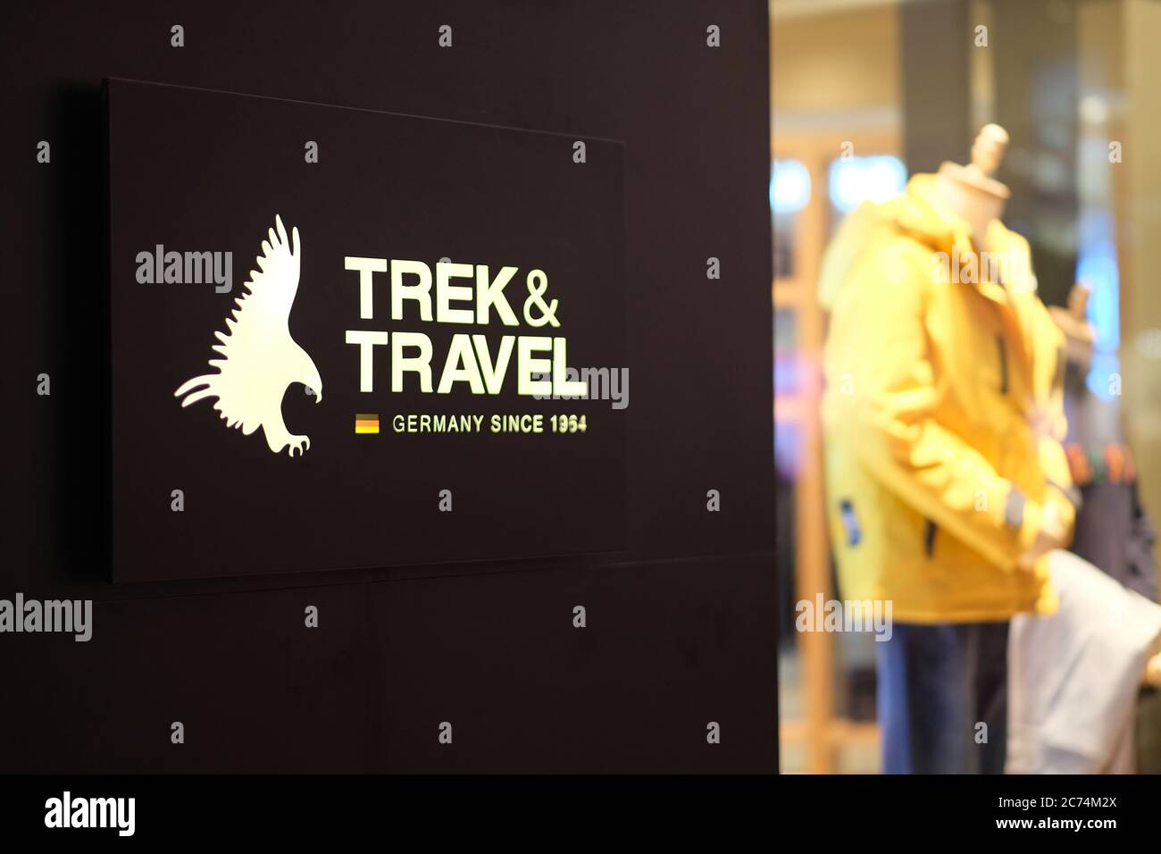 Primo piano il logo Trek&Travel illuminato sulla parete scura. Sfocatura abbigliamento vetrina negozio come sfondo. Un marchio di abbigliamento tedesco da uomo Foto Stock