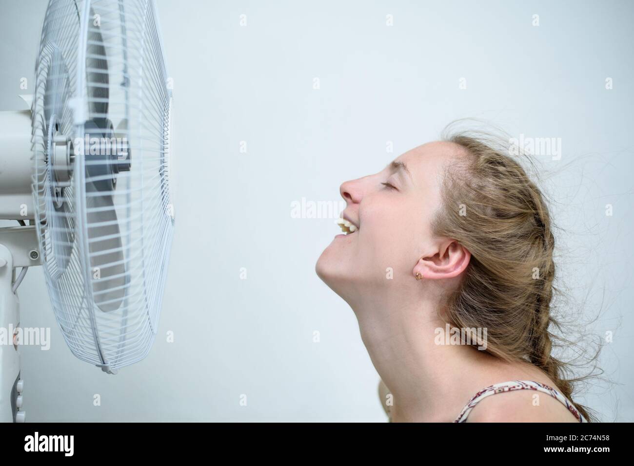 Giovane donna seduta davanti al ventilatore elettrico che si sente calda  nell'appartamento e cerca di catturare l'aria fredda