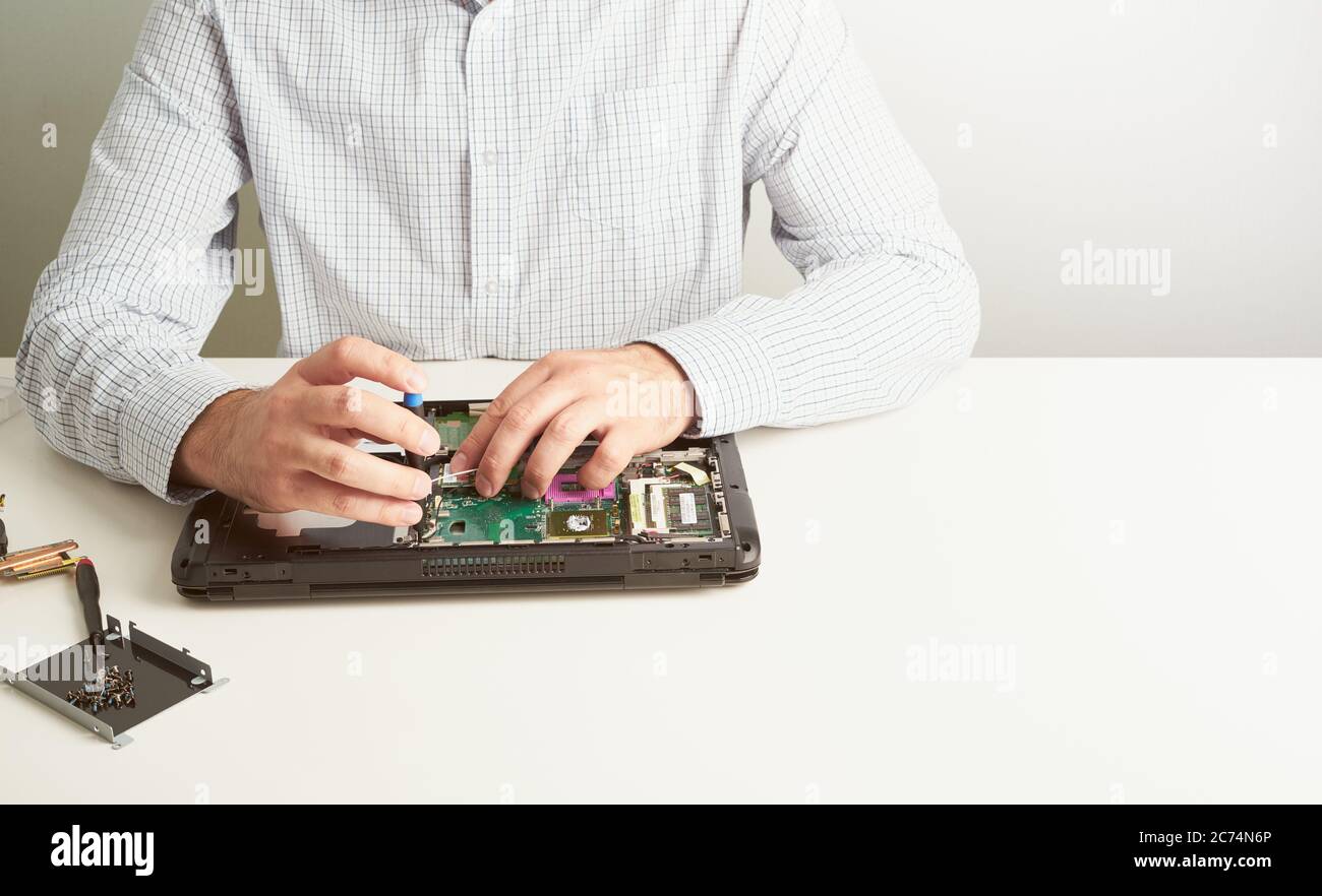 Uomo ripara il computer. Un tecnico dell'assistenza in camicia ripara il laptop, presso la scrivania bianca contro la parete bianca. Foto Stock