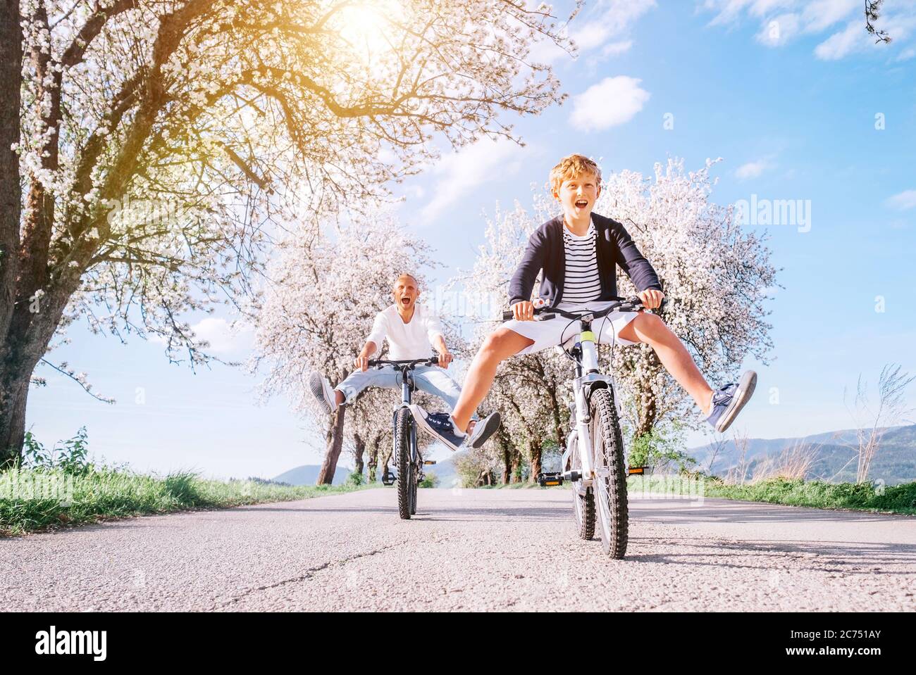 Padre e figlio si divertono a spargere gambe larghe e a urlare quando si cavalcano le biciclette su strada di campagna sotto alberi fioriti. Concetto di stile di vita sportivo sano Foto Stock