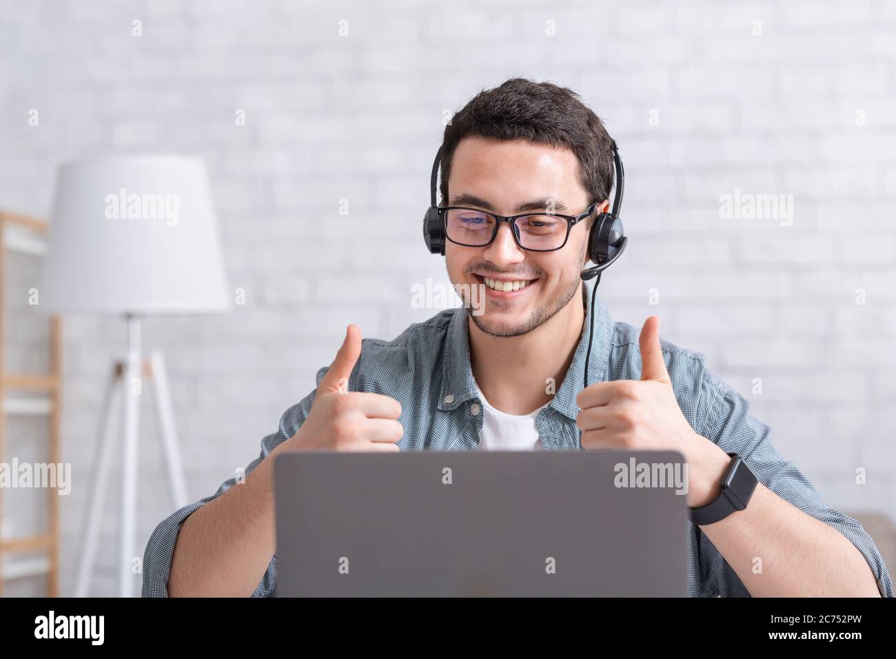 Un ragazzo sorridente con gli occhiali punta le dita verso l'alto e guarda il computer portatile nell'interno del salotto Foto Stock