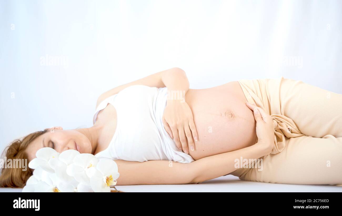 Donna incinta praticando esercizi prenatali respiratori con belle orchidee fiori su sfondo bianco. Concetto di relax e preparazione alla consegna Foto Stock