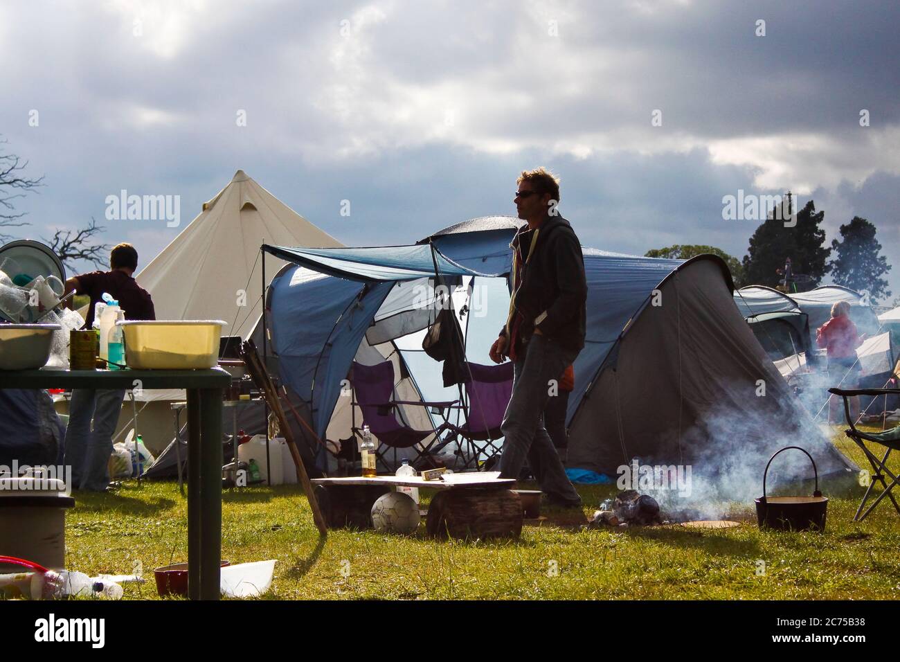 Preparandosi a cucinare su un falò in un campeggio festival con le nuvole di pioggia buie e il sole, Norfolk Foto Stock