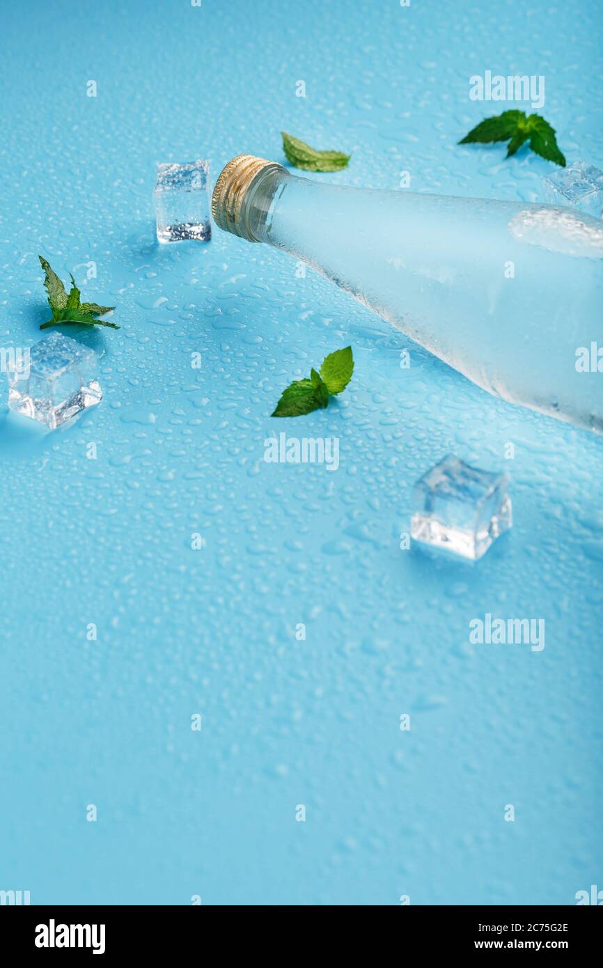 Una bottiglia di acqua ghiacciata, cubetti di ghiaccio, gocce e foglie di menta su sfondo blu. Foto Stock