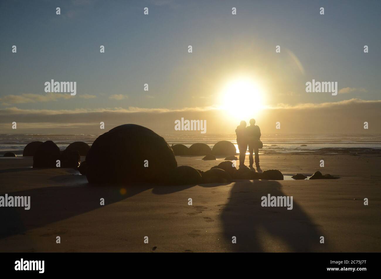 Silhouette di coppie che scattano foto a Moeraki Boulders durante il tramonto, con l'angelo proiezione ombra sulla sabbia. Foto Stock