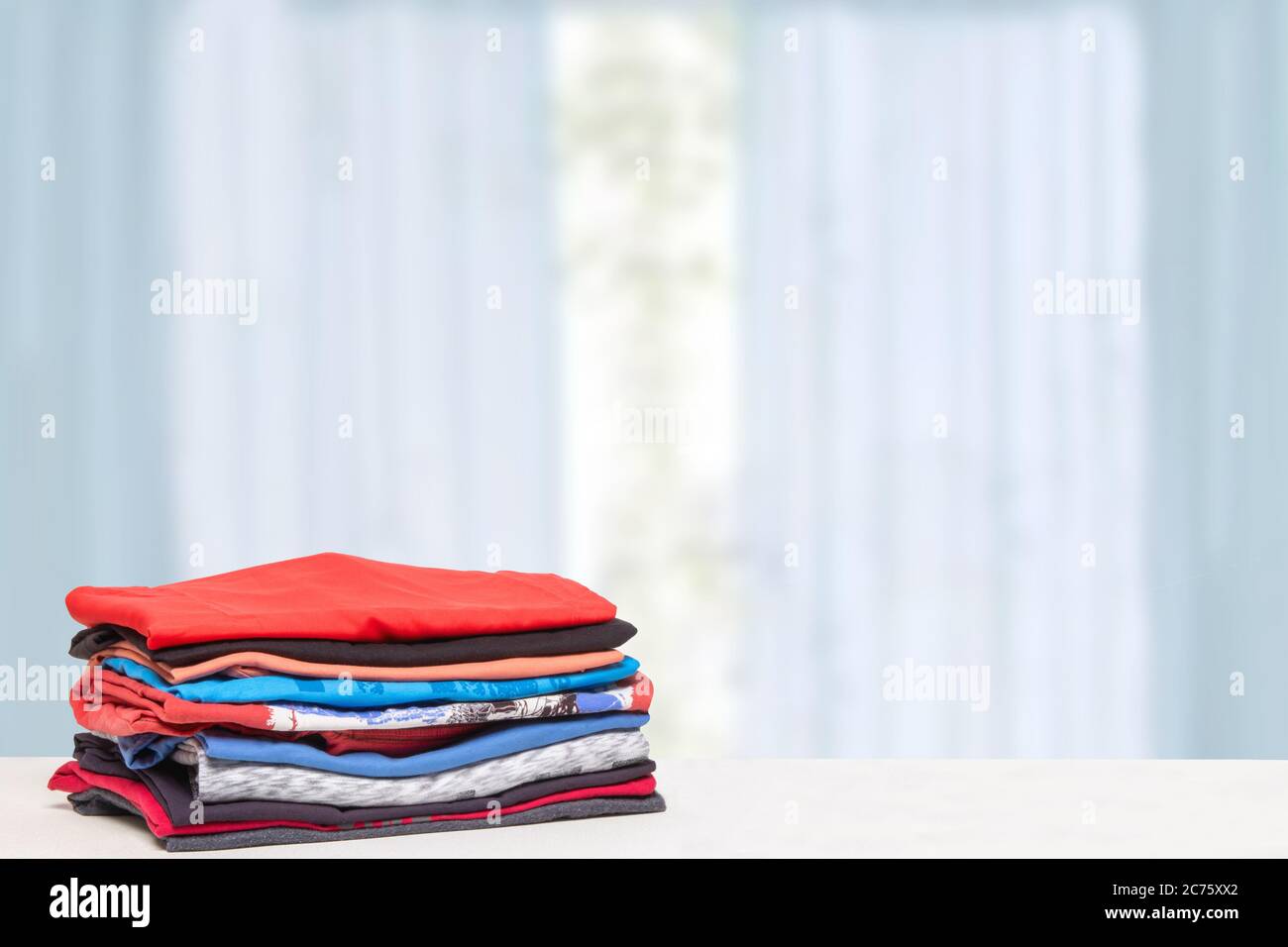 Accatastare abiti colorati. Pile di t-shirt o camicie in cotone ripiegato su un tavolo luminoso con spazio per il montaggio del prodotto su tende sfocate b Foto Stock