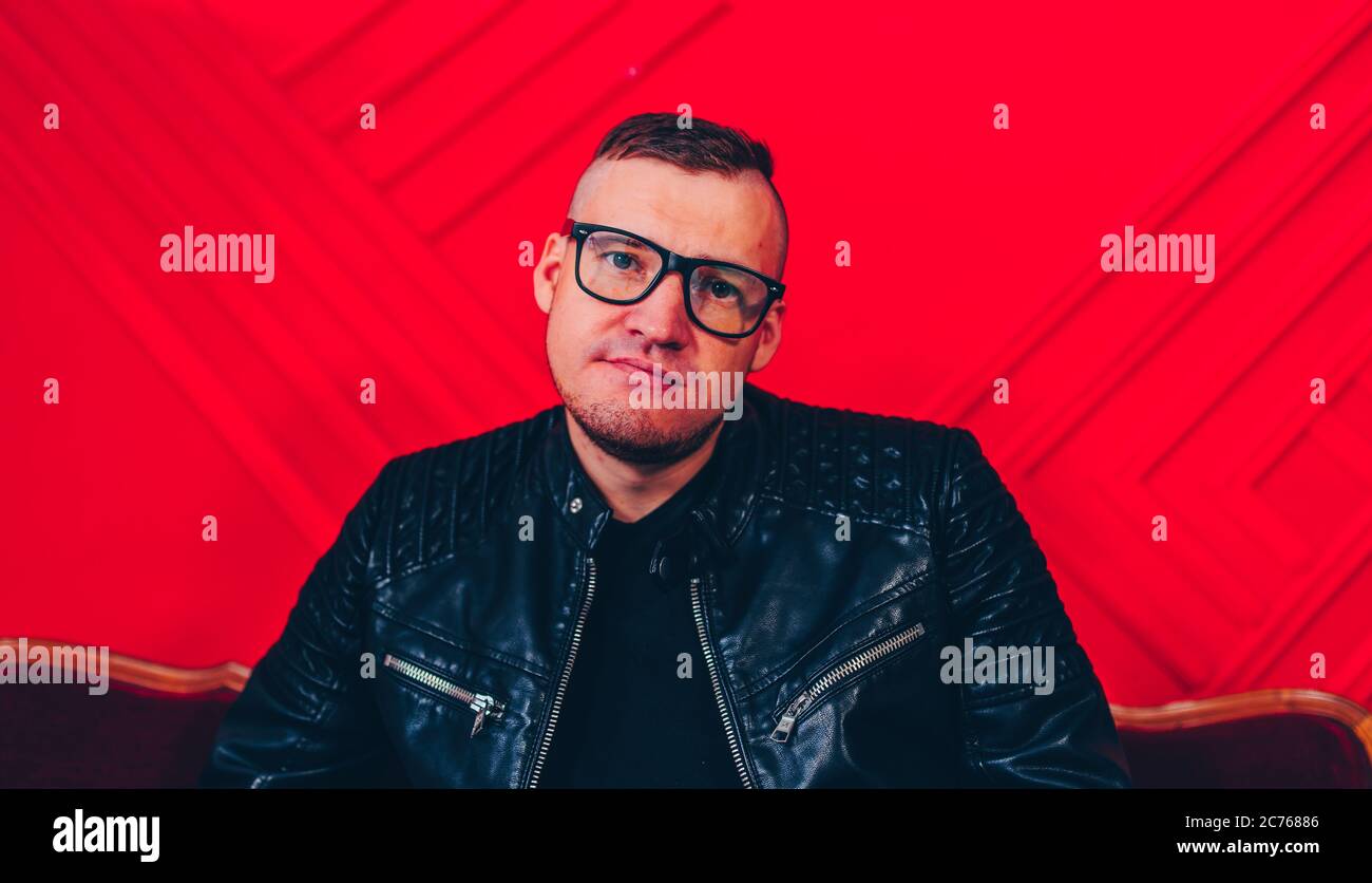 Bel giovanotto con bicchieri poggiati su un piccolo divano. Uomo di charme adulto in posa, seduta su poltrona contro parete rossa. Foto Stock