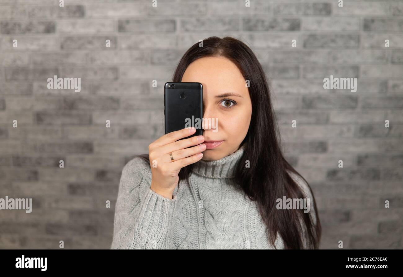 Una giovane bella donna chiude l'occhio con un telefono. La donna si nasconde dietro un telefono cellulare. Il concetto di guardare attraverso il prisma dello schermo Foto Stock