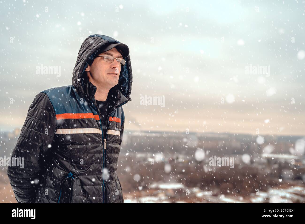 Uomo adulto in serata innevata. Uomo adulto in outwear con cappuccio e occhiali in piedi sotto la neve caduta contro la finestra del tramonto nella campagna invernale Foto Stock