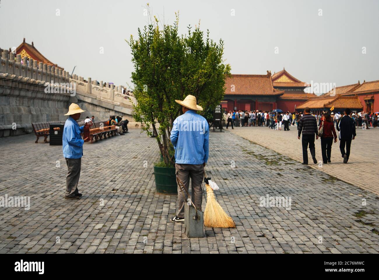 Due addetti alla manutenzione che hanno una conversazione nella zona della Città Proibita, indossando cappelli di paglia e magliette blu, turisti in background. Foto Stock