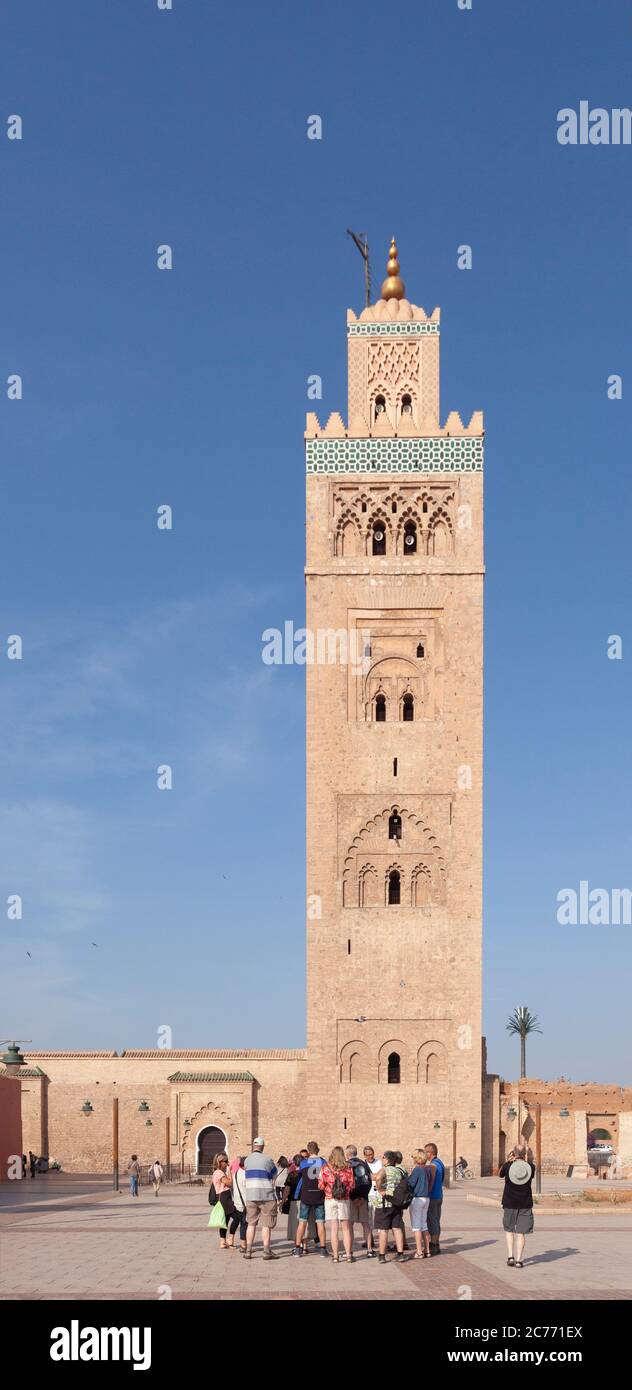 Un gruppo di turisti in un tour guidato che si trova fuori dalla Moschea di Kutubiyya nel centro di Marrakech. Il fusto quadrato del suo Minar in arenaria in stile Almohad Foto Stock