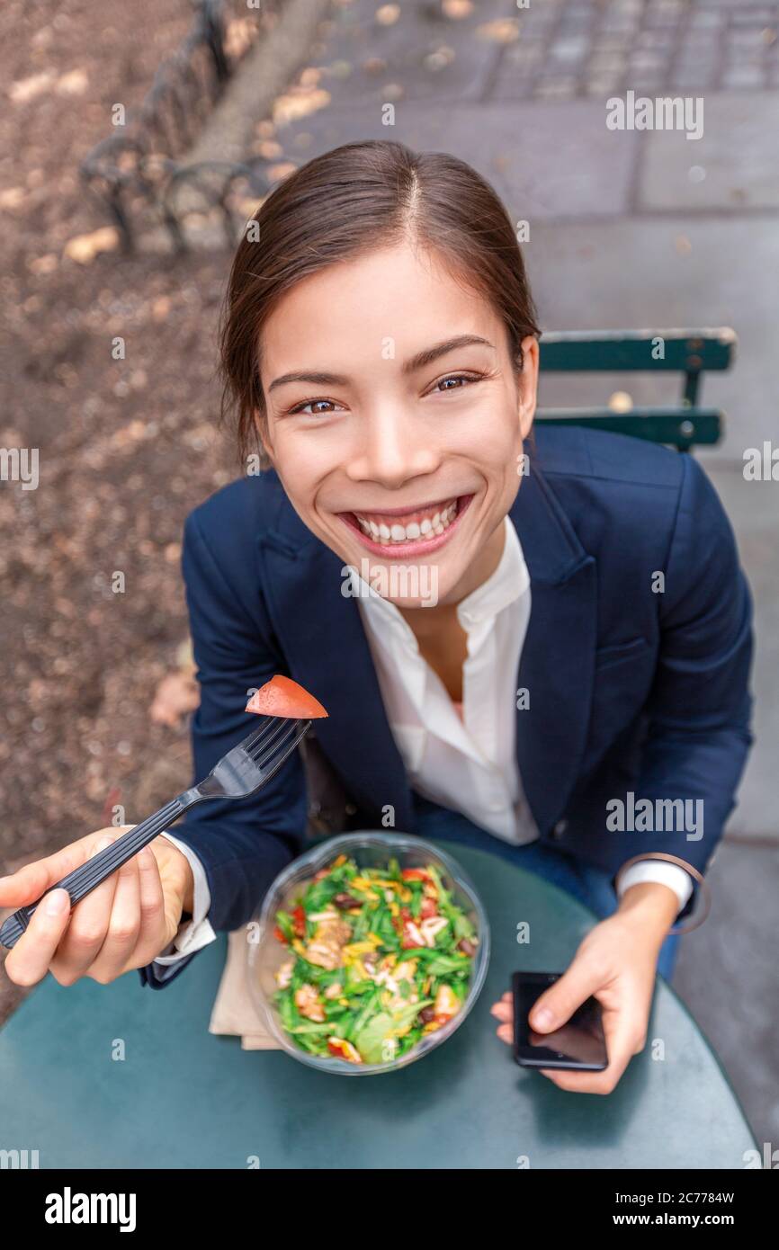 Pausa pranzo sano mangiare asiatico donna d'affari pronto a mangiare insalata ciotola nel parco della città vivere stile di vita urbano utilizzando il telefono. Felice sorridente multirazziale Foto Stock