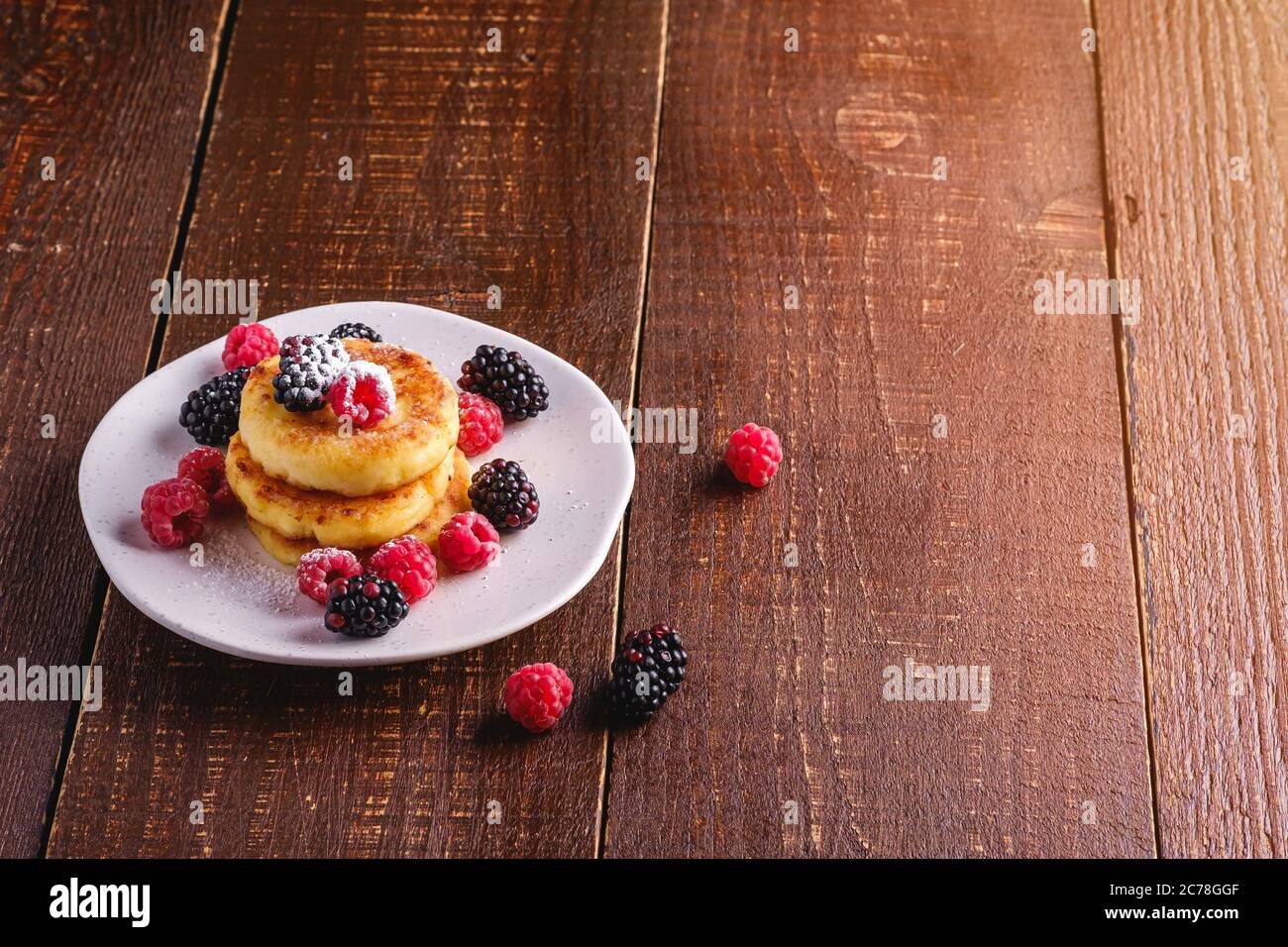 Frittelle di formaggio e zucchero in polvere, frittelle di cagliata dessert con lampone e frutti di bosco in piatto su sfondo di legno marrone scuro Foto Stock