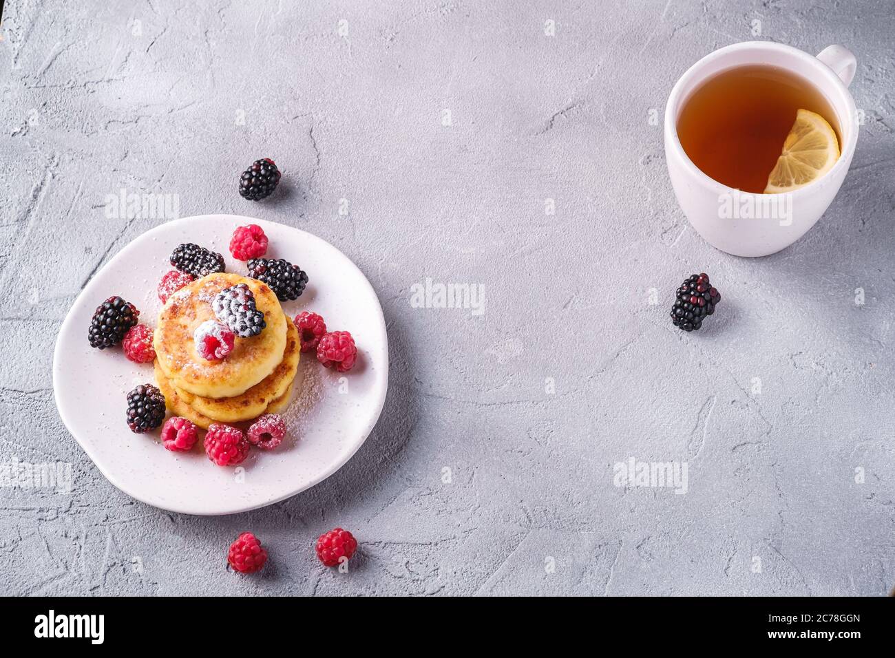 Frittelle al formaggio e zucchero in polvere, frittelle di cagliata dessert con lampone e frutti di bosco in piatto vicino alla tazza di tè caldo con fetta di limone Foto Stock