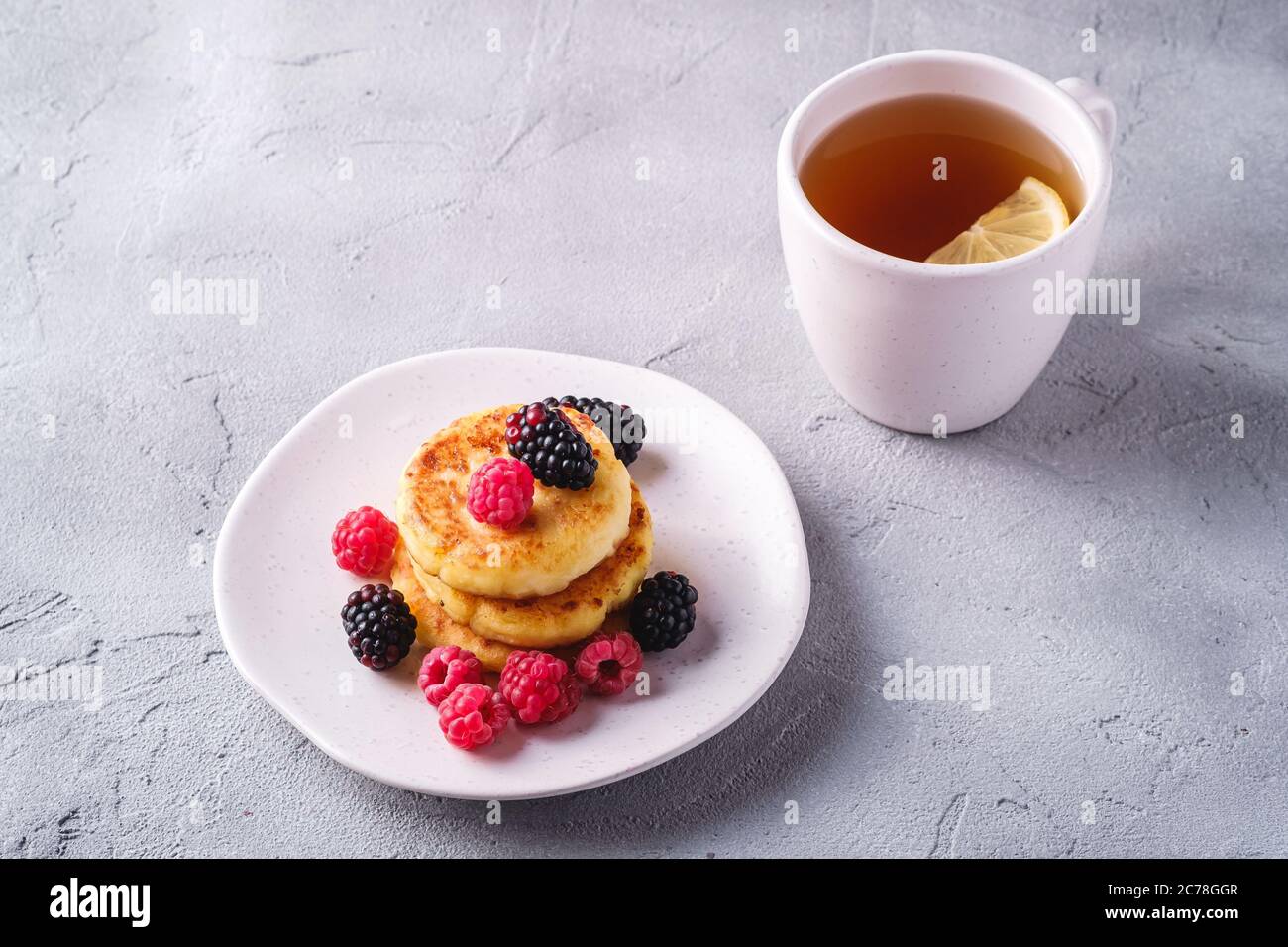 Frittelle di formaggio cottage, frittelle di cagliata dessert con lampone e frutti di bosco in piatto vicino alla tazza di tè caldo con fetta di limone Foto Stock