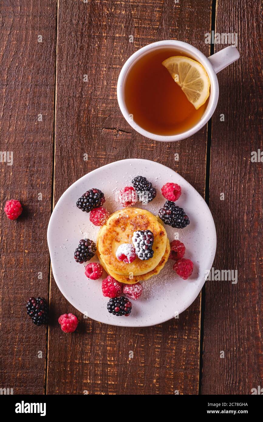 Frittelle di formaggio cottage, frittelle di cagliata dessert con lampone e frutti di bosco in piatto vicino alla tazza di tè caldo con fetta di limone Foto Stock