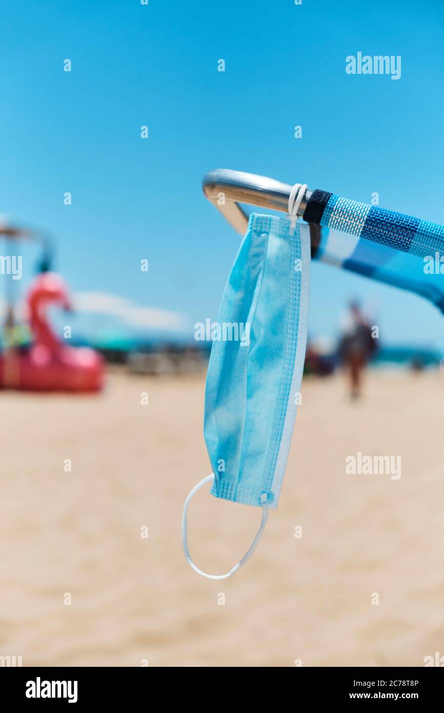 primo piano di una maschera chirurgica appesa su una sedia blu sulla spiaggia, con il mare sullo sfondo Foto Stock