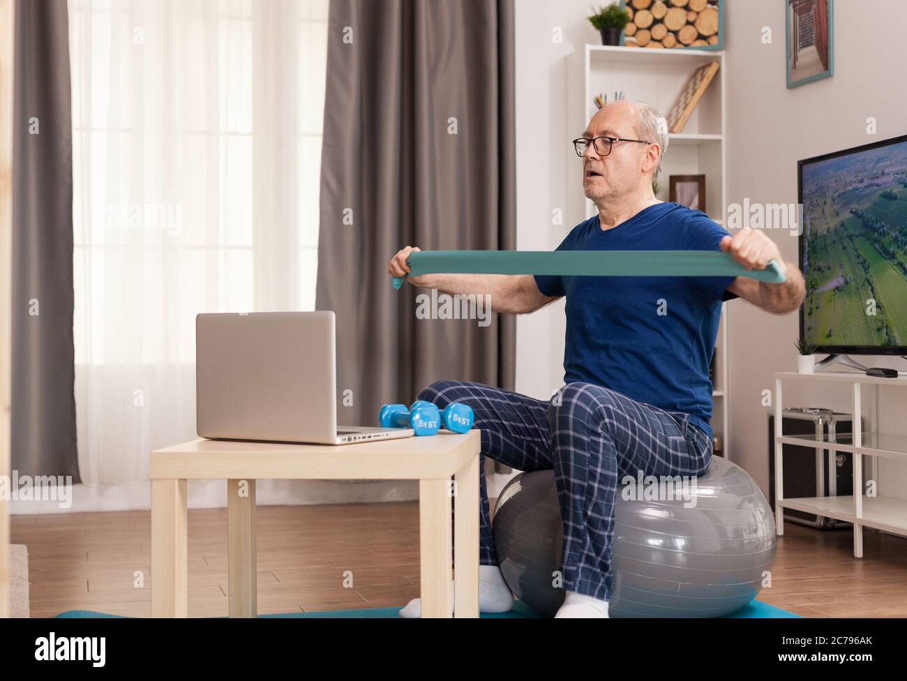 Forte allenamento online con la banda di resistenza. Anziano pensionato online internet esercizio di formazione a casa attività sportiva con manubri, banda di resistenza, palla svizzera all'età di anziano pensionamento. Foto Stock