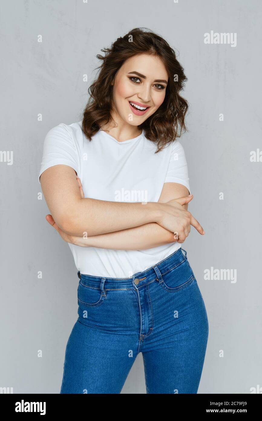 Ragazza positiva in jeans, maglietta bianca e paletti rossi che si posano vicino alla parete tenendo le mani incrociate sul petto Foto Stock