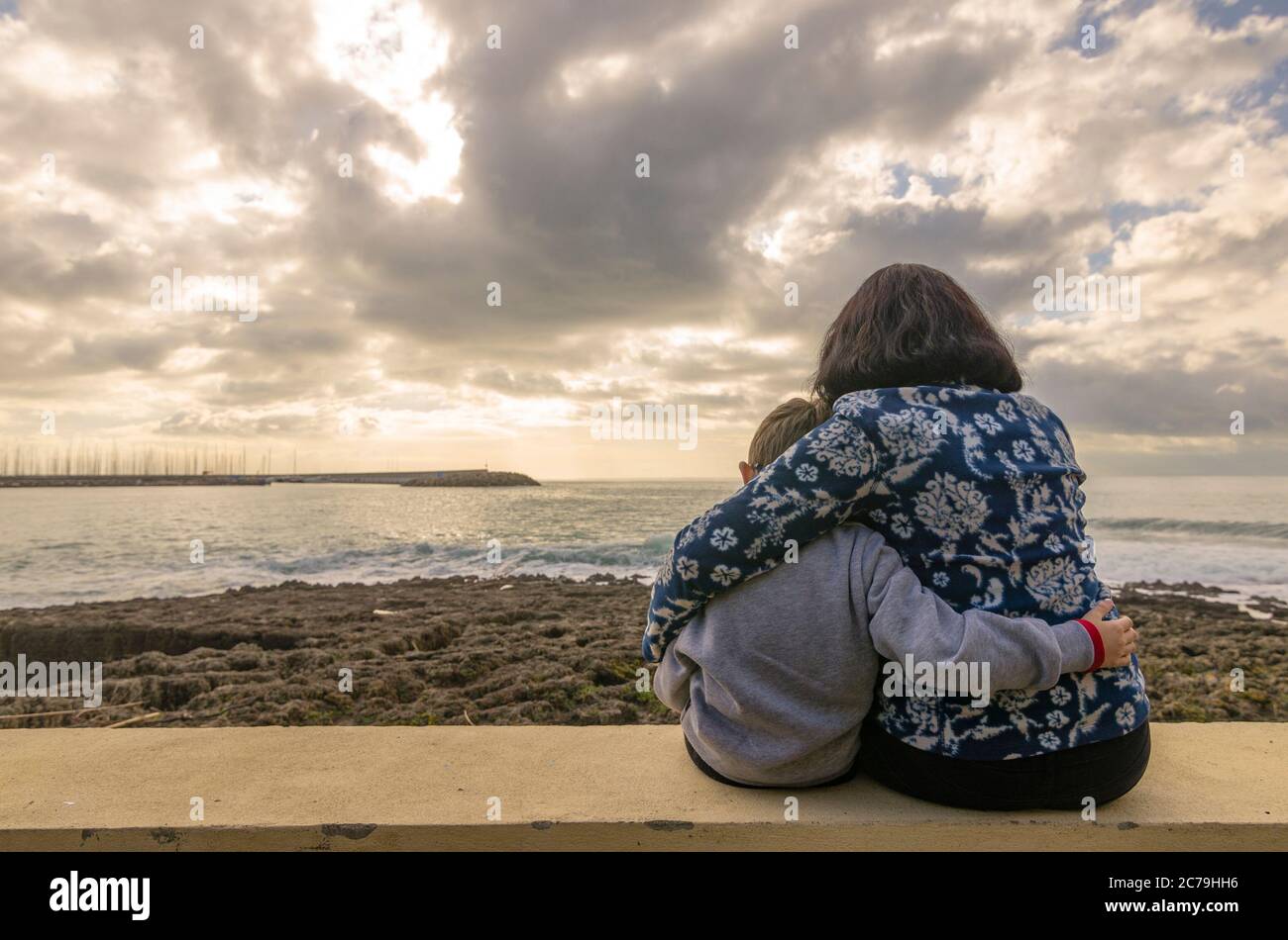 Mamma con figlio seduto e abbracciato l'un l'altro sul muro che guarda al mare Foto Stock