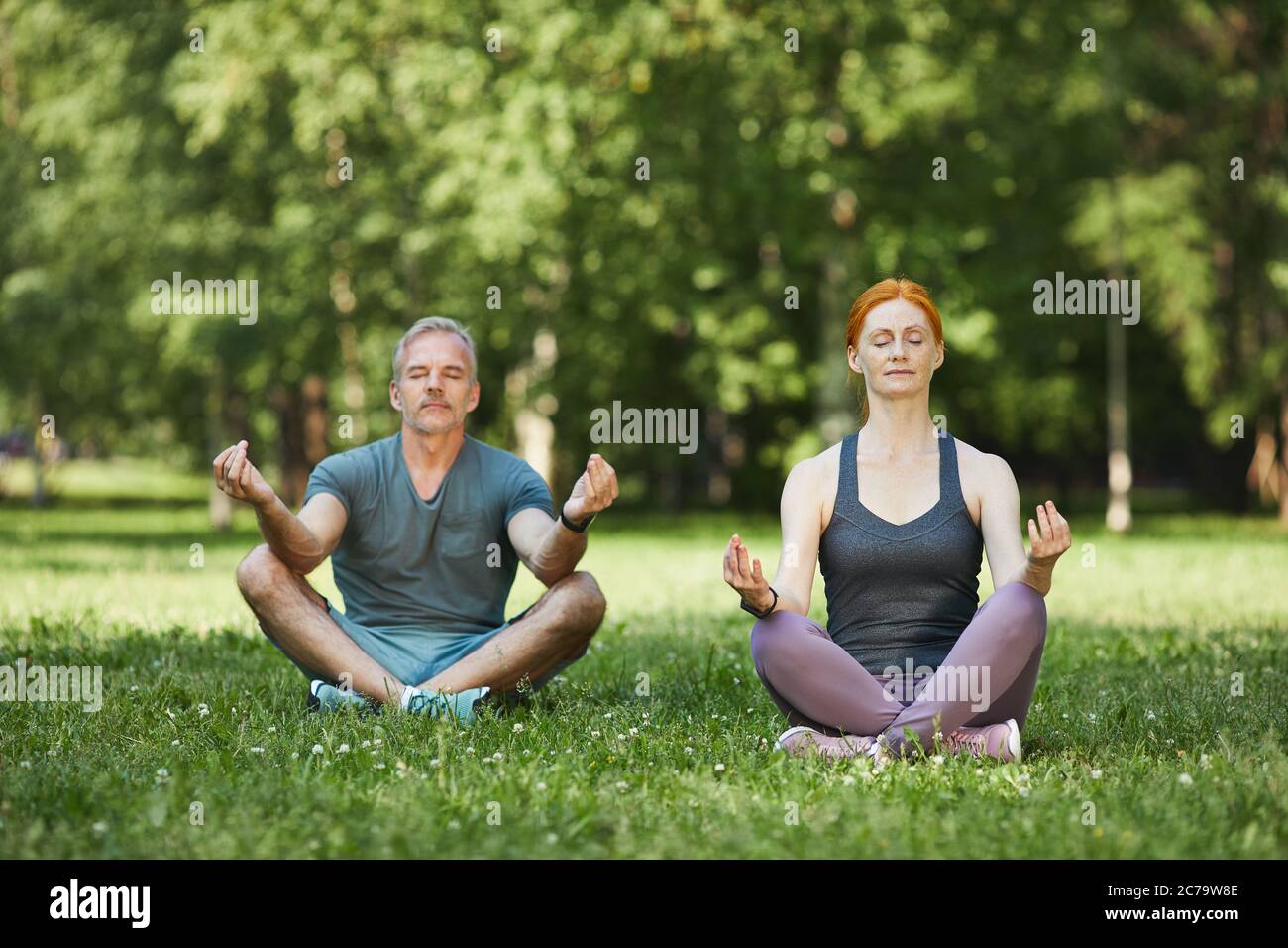 Coppia matura e tranquilla concentrata sulla mente durante la meditazione seduta con gambe incrociate in un bellissimo parco Foto Stock