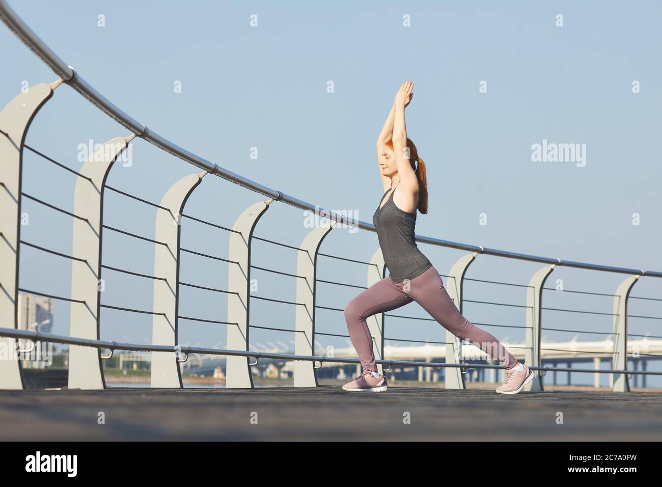 Donna di mezza età sportiva con occhi chiusi che eseguono la posizione guerriero con braccia sollevate sull'argine della città Foto Stock