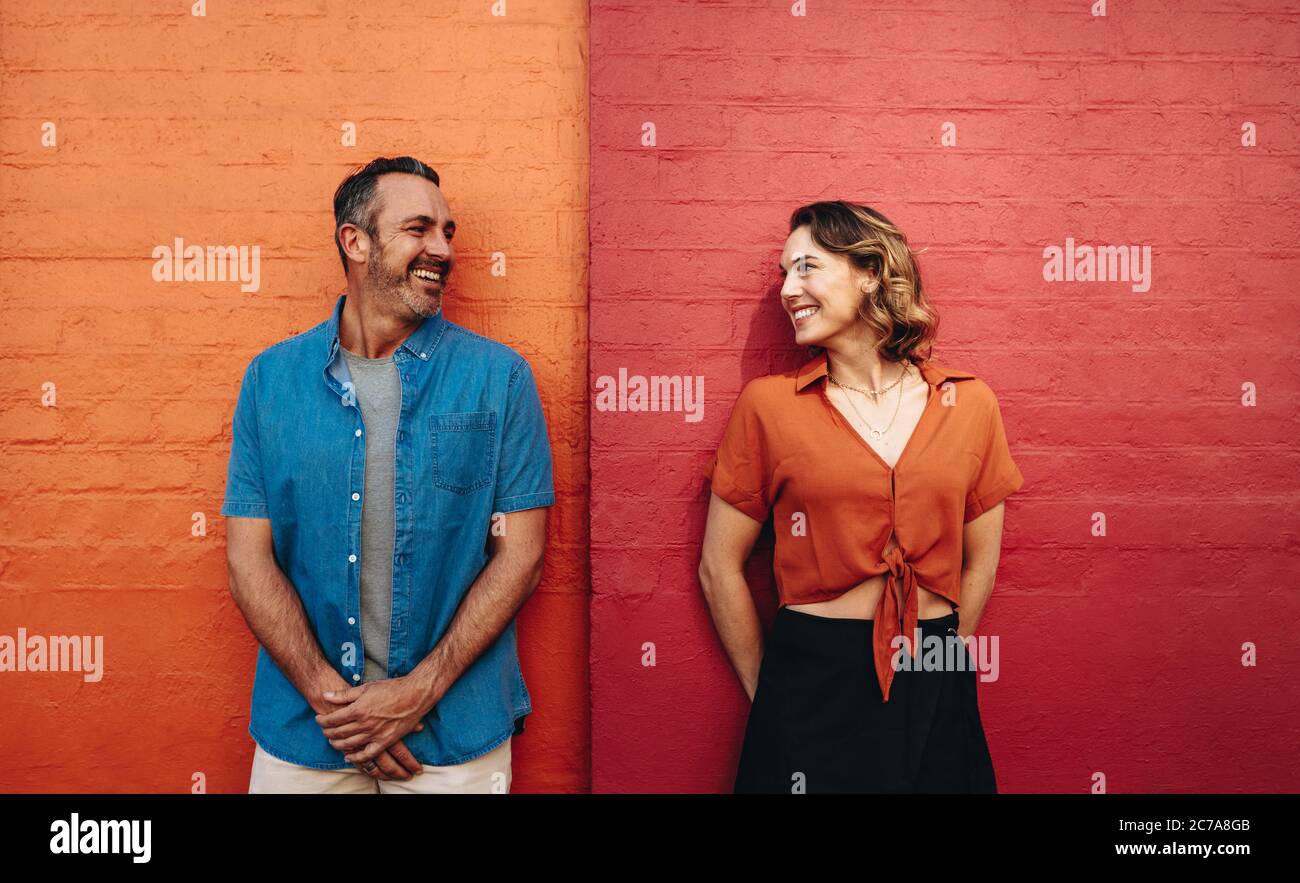 Uomo e donna in piedi da un muro che si guarda l'un l'altro e sorridente. Coppia appoggiata a una parete di colore rosso e arancione. Foto Stock