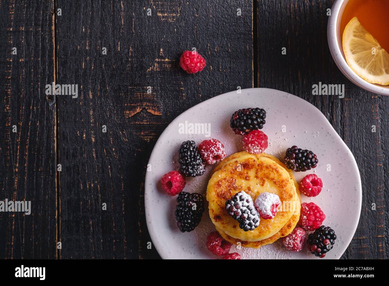 Frittelle di formaggio cottage, frittelle di cagliata dessert con lampone e frutti di bosco in piatto vicino alla tazza di tè caldo con fetta di limone Foto Stock